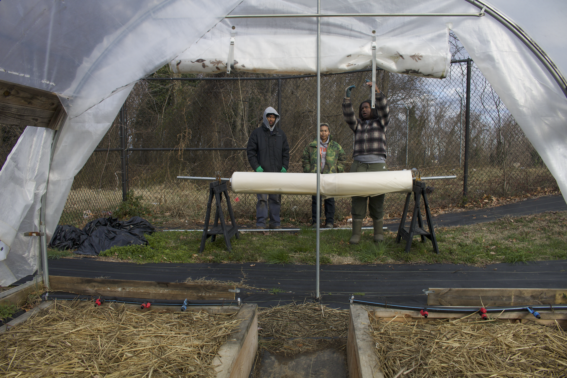 Three people standing in front of a hoop house, one person explaining something to them as though they are preparing to work